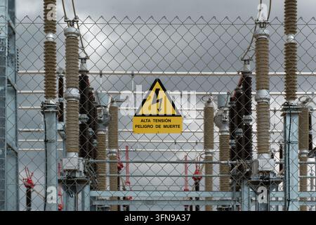 Gelbes dreieckiges Hochspannungsgefahrenschild an einem Drahtgitterzaun, der eine elektrische Unterstation mit keramischen Isolatoren umgibt. Stockfoto