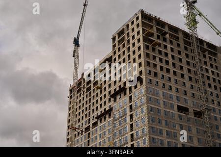 Graue Wolken schweben über einem Betonhochhaus mit freiliegendem Stahlgerüst und unfertigen Stockwerken. Große Turmdrehkräne und umlaufende Gerüste rahmen die Gebäudefassade ein Stockfoto