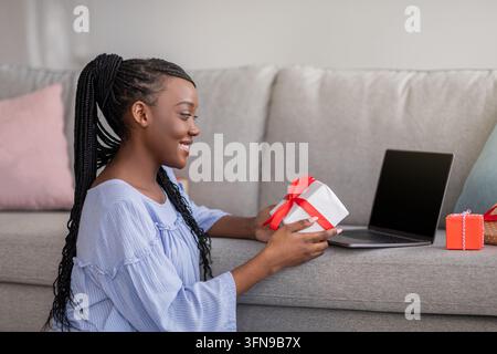 Lächelnde junge schwarze Frau mit Dreadlocks, die ein Geschenk hält, Laptop mit leerem Bildschirm benutzt, Seitenansicht, Kopierraum. Hübsche afrikanische Dame, die ein Geschenk öffnet, ha Stockfoto