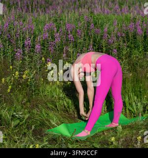 Sportliche Frau in rosa Sportkleidung macht eine stehende Vorwärtsbeuge auf einer grünen Matte in einer blühenden Wiese. Outdoor-Yoga- und Pilates-Training zwischen Wildblumen Stockfoto