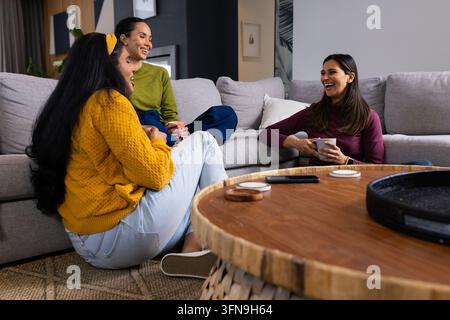 Verschiedene Freundinnen, die auf einem grauen Sofa und dem Boden um einen hölzernen Couchtisch sitzen, Becher in der Hand. Gemütlich, Lounge, Gespräch, Frauen, Tassen, Dekor, warm Stockfoto