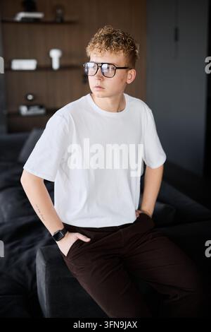 Junger weißer Mann steht an einem Regal, Brille und gelassene Haltung, überprüft mentale Notizen vor dem Treffen, weiches Tageslicht, minimalistisch Stockfoto