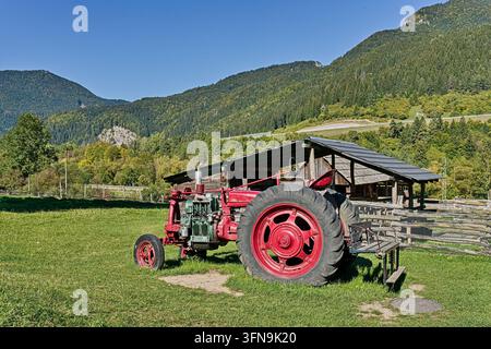 Roter Vintage-Bauernhofstraktor, der auf einer Wiese in der Nähe eines Schuppens mit Bergkamm im Hintergrund geparkt ist. Stockfoto