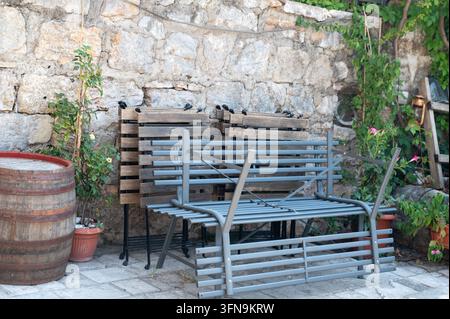 Gestapelte Metall-Stühle im Freien lehnen an einer sonnenbeschienenen Steinmauer in einem rustikalen Innenhof mit Topfpflanzen und Holzfass sowie Leiter für Garten oder Café Stockfoto