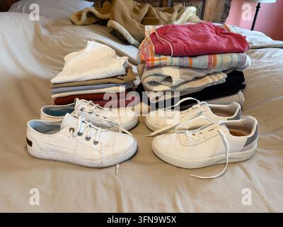 Zwei Stapel ordentlich gefalteter Kleidung in Weiß, Grau, Rosa und karierten Mustern stehen neben zwei Paar weißen Canvas-Sneakern, die als Teil des auf einem Bett arrangiert sind Stockfoto