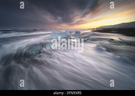 Blick auf Diamond Beach mit glitzernden Eisbrocken, die über den schwarzen Sandstrand verstreut sind, während Wellen unter einem dramatischen Sonnenuntergangshimmel über sie hinwegspülen in Sveitarf Stockfoto