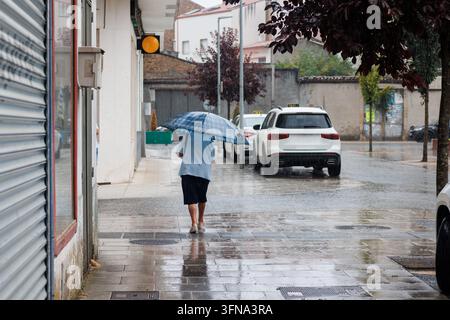 Eine ältere Dame mit einem blau karierten Regenschirm geht an einem nassen Gehweg an einem regnerischen Tag in Melide, Galicien, Spanien, mit Autos im Hintergrund. Stockfoto