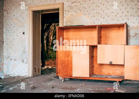 Mid-Century-Holzkabinett in einem verlassenen Raum mit abblätternder Tapete und einer offenen Tür nach draußen. Selektive Fokussierung. Stockfoto