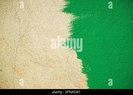 Lebendige grüne Farbe bedeckt teilweise eine raue, beige verputzte Wand mit sichtbaren Pinselstrichkanten und Oberflächenstruktur. Stockfoto