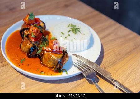 Türkisch-stilvolle Auberginen-Wrap-Mehlspeisen serviert mit Tomatensauce, weißem Reis und Kirschtomaten auf einem weißen Teller auf einem Holztisch. Stockfoto