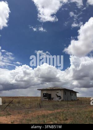 Verfallenes, verlassenes Haus auf einem ländlichen Feld unter einem teilweise bewölkten Himmel, umgeben von trockenem Gras und rustikalen Zäunen in New Mexico Stockfoto