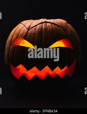 Eine Nahaufnahme im Studio eines bedrohlichen Jack-o'-Lantern, der mit scharfen, schrägen Augen und einem gezackten Mund geschnitzt ist und mit einem intensiven orangefarbenen Licht leuchtet. Stockfoto