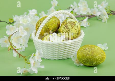 Marmorierte grüne Ostereier in einem weißen geflochtenen Korb mit Frühlingsblüten auf grünem Hintergrund. Ökofreundliches Feiertagskonzept, Nahaufnahme Stockfoto