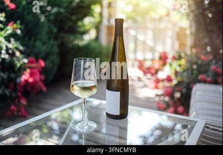Glas weißer Wein neben einer Flasche auf einem Glastisch in einem Garten voller lebendiger Blumen und üppiger Grünpflanzen im Sonnenlicht Stockfoto