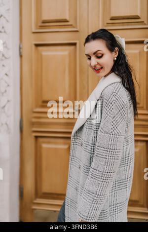 Damenmode-Mantel, der in einem stilvollen grauen Karo-Outfit lächelt, während sie vor einer großen Holztür steht, mit Platz für Text Stockfoto