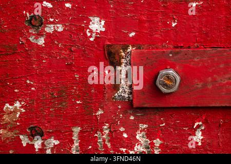 Nahaufnahme-Hintergrund einer verwitterten und abblätternden rot gestrichenen Holzoberfläche, mit einer rustikalen roten Metallstange, die mit einem rostigen Metallbolzen befestigt ist und ol Stockfoto