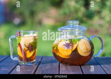 Gesunder Apfeltee in einer Glas-Teekocher und Becher im Sommergarten auf Holztisch. Nahaufnahme von Apfeltee mit Zimt, Nelken oder Minzblättern über die Natur b Stockfoto