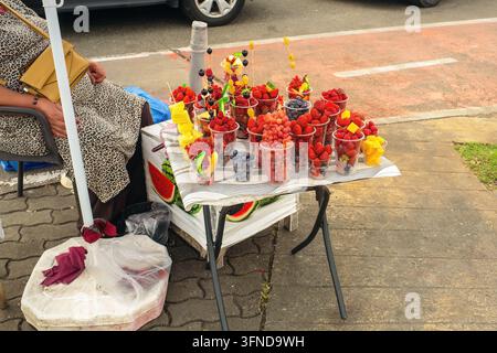 Frische Beeren und geschnittenes Obst in Plastikbechern auf dem Tisch eines Straßenhändlers auf dem Bürgersteig einer Stadt, kleines Geschäft in einer Ferienstadt. Stockfoto