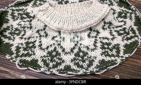 Ein weiß-grünes, handgestricktes Rundkragen mit einem Jacquardmuster liegt auf einem dunklen Holztisch neben dekorativen Blumen. Detail eines zukünftigen Pullovers. Stockfoto