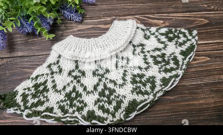 Ein weiß-grünes, handgestricktes Rundkragen mit einem Jacquardmuster liegt auf einem dunklen Holztisch neben dekorativen Blumen. Detail eines zukünftigen Pullovers. Stockfoto