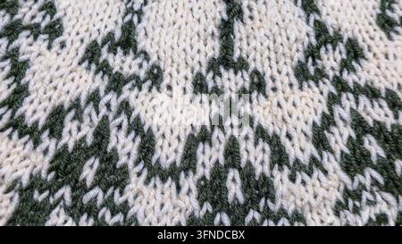 Ein weiß-grünes, handgestricktes Rundkragen mit einem Jacquardmuster liegt auf einem dunklen Holztisch neben dekorativen Blumen. Detail eines zukünftigen Pullovers. Stockfoto