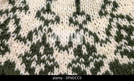 Ein weiß-grünes, handgestricktes Rundkragen mit einem Jacquardmuster liegt auf einem dunklen Holztisch neben dekorativen Blumen. Detail eines zukünftigen Pullovers. Stockfoto