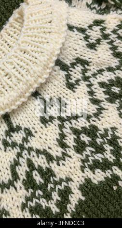 Ein weiß-grünes, handgestricktes Rundkragen mit einem Jacquardmuster liegt auf einem dunklen Holztisch neben dekorativen Blumen. Detail eines zukünftigen Pullovers. Stockfoto