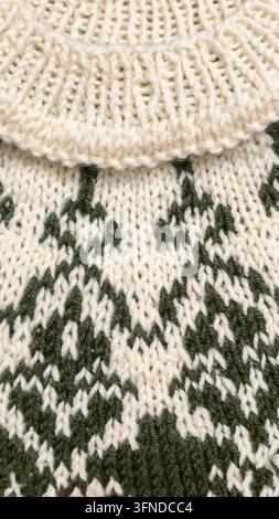 Ein weiß-grünes, handgestricktes Rundkragen mit einem Jacquardmuster liegt auf einem dunklen Holztisch neben dekorativen Blumen. Detail eines zukünftigen Pullovers. Stockfoto
