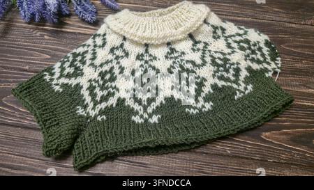 Ein weiß-grünes, handgestricktes Rundkragen mit einem Jacquardmuster liegt auf einem dunklen Holztisch neben dekorativen Blumen. Detail eines zukünftigen Pullovers. Stockfoto