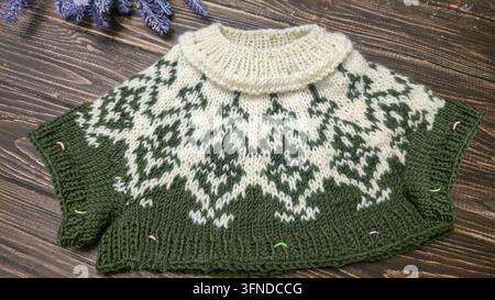 Ein weiß-grünes, handgestricktes Rundkragen mit einem Jacquardmuster liegt auf einem dunklen Holztisch neben dekorativen Blumen. Detail eines zukünftigen Pullovers. Stockfoto