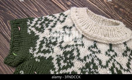 Ein weiß-grünes, handgestricktes Rundkragen mit einem Jacquardmuster liegt auf einem dunklen Holztisch neben dekorativen Blumen. Detail eines zukünftigen Pullovers. Stockfoto