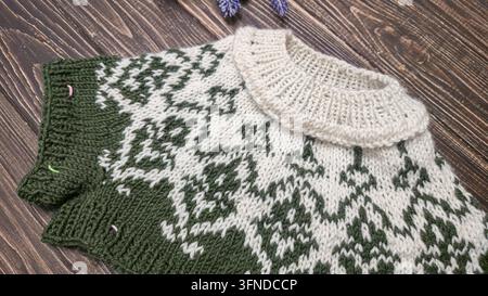 Ein weiß-grünes, handgestricktes Rundkragen mit einem Jacquardmuster liegt auf einem dunklen Holztisch neben dekorativen Blumen. Detail eines zukünftigen Pullovers. Stockfoto