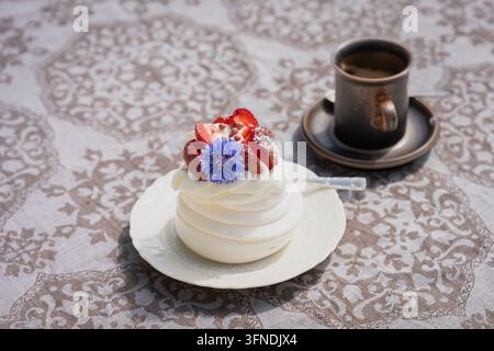 Pavlova-Dessert mit Schlagsahne, frischen Erdbeeren und einer lebhaften blauen Kornblume, serviert auf einem eleganten weißen Teller mit einer Tasse Kaffee. Stockfoto