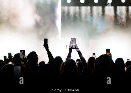 Silhouetten von Händen und Mobiltelefonen, die in der Luft erhoben sind, um eine helle Live-Show auf der Bühne mit Nebel und Bühnenlichtern aufzunehmen. Stockfoto