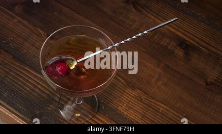 Eine Nahaufnahme eines Manhattan-Cocktails in einem Coupé-Glas, garniert mit einer Kirsche und einem Barlöffel, der darin ruht, vor einem dunklen Holztisch. Stockfoto