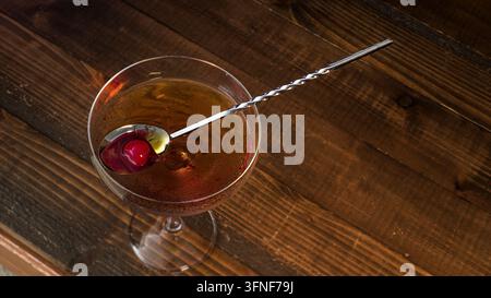 Manhattan-Cocktail, garniert mit einer Kirsche, ruht in einem Coupé-Glas, mit einem langen Barlöffel darin, präsentiert auf einer strukturierten Holzoberfläche bei einem b Stockfoto