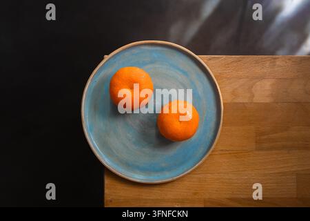 Überkopf-Stillleben mit zwei Klementinen auf einem handglasierten blauen Keramikteller, der auf der Ecke eines Holztisches platziert ist, vor dunklem Hintergrund. Stockfoto