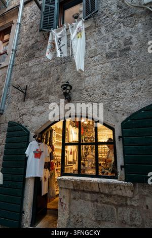 Kotor, Montenegro - 6. September 2026: Ein kleiner Souvenirladen befindet sich in einem gewölbten Steinladen mit grünen Holzläden und Wäsche darüber in Ko Stockfoto