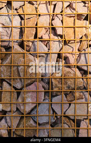 Nahaufnahme eines gelben Metallgitterzauns, der eine strukturierte Gabionwand aus bunten Steinen hält Stockfoto