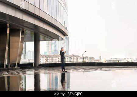 Vollständige Figur einer professionellen Geschäftsfrau, die ein Smartphone auf einer modernen Büroterrasse mit einer Stadtsilhouette und Wasserreflexion im Hintergrund benutzt Stockfoto