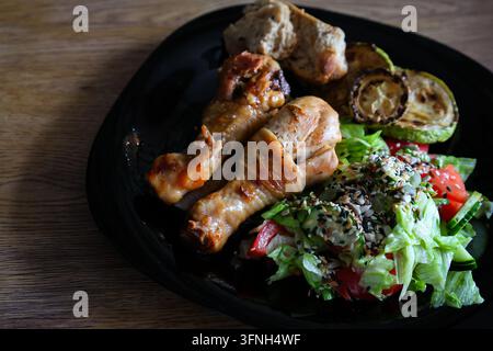 Gebratene Hähnchen-Schenkel mit frischem Salat und Brot auf schwarzem Teller. Gegrilltes Hähnchen mit gebratenem Zucchini und Salat mit Samen. Hausgemachte Speisen serviert. Stockfoto