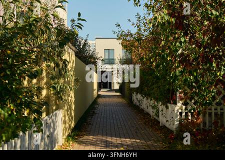 Perspektivische Aufnahme einer gepflasterten Gasse, die von Holzzäunen und üppigem grünen Laubwerk gesäumt ist und an einem sonnigen Tag auf ein modernes helles Stadthaus zuführt. Stockfoto