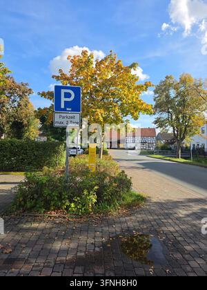 Blaues Parkverkehrsschild mit einer Beschlagstafel für Beschränkungen, montiert auf einem Metallpfahl in einem Parkplatzbereich mit Herbstbäumen und einem traditionellen deutschen Haus Stockfoto