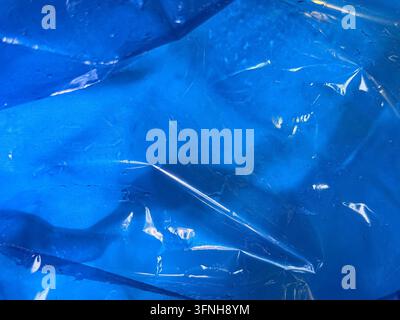 Nahaufnahme von lebhaftem blauem Stoff, der von einer durchsichtigen, zerknitterten Plastikfolie bedeckt ist. Licht funkelt auf der Oberfläche und wird von subtilen Wassertropfen reflektiert, die Stockfoto