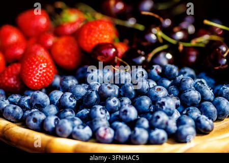 Makroaufnahme von reifen süßen Blaubeeren, frischen Erdbeeren und dunklen Kirschen, serviert auf einem natürlichen Holzteller. Stockfoto