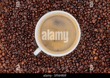 Oberblick auf eine weiße Espressotasse mit reicher Crema, umgeben von gerösteten Kaffeebohnen, auf dunklem Steingut-Hintergrund für Café-Werbung Stockfoto