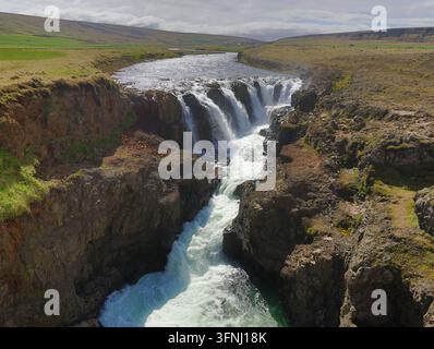 Mächtiger Sjonarhorn-Wasserfall, der durch eine tiefe Felsschlucht in einem weiten grünen Tal unter einer bewölkten Himmeldecke rauscht, von oben in einem wilden Norden aufgenommen Stockfoto