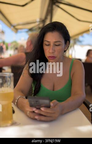 Eine Frau in einem grünen Oberteil nutzt ihr Smartphone, während sie an einem Außentisch eines Cafés mit einem Bierglas sitzt. Stockfoto