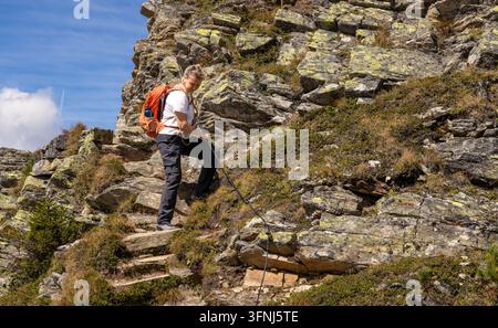 Wandererin steigt einen steilen, felsigen Bergpfad mit einem orangefarbenen Rucksack hinauf und benutzt einen Wanderstock an einem sonnigen Tag unter blauem Himmel und vereinzelten Wolken. Stockfoto