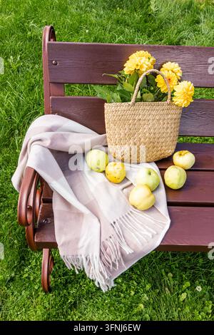 Ein geflochtener Korb gefüllt mit gelben Blumen steht auf einer hölzernen Parkbank neben frischem Apfel und einer gefransten Decke und schafft so eine fröhliche Sommerpicknick-Szene Stockfoto